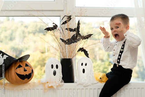 Halloween. Concept. A cute and cheerful boy sits near the window against the background of white ghosts and a vase with dry branches and black paper bats.