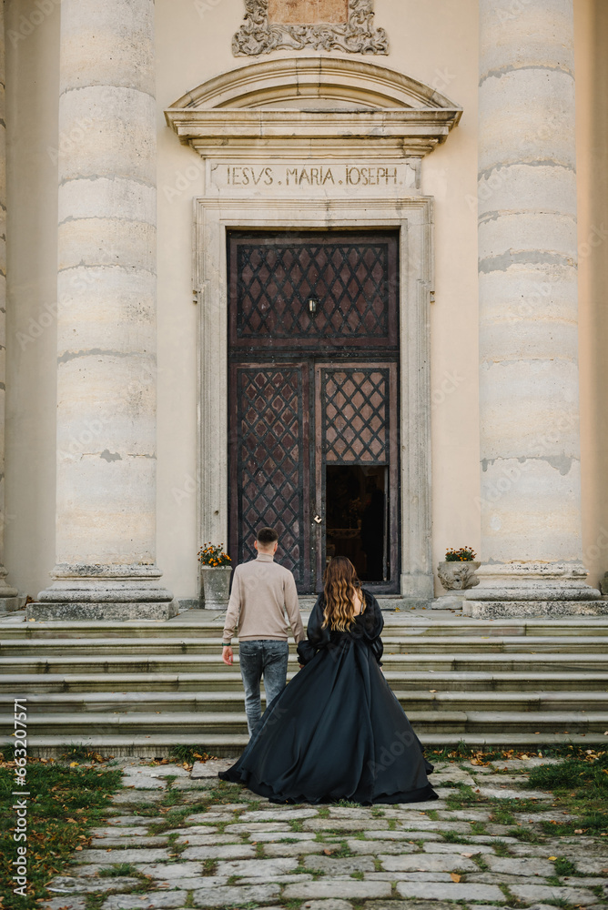 Newlyweds walk near Baroque Roman Catholic church in Pidhirtsi Ukraine. Bride and groom walking on stairs near large columns of ancient temple at sunset. Couple hold hands nature autumn day. Back view