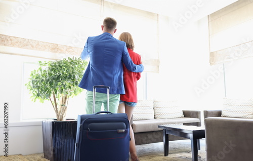 Portrait of couple dressed in suits, suitcase behind, enjoy view out of window, wait for flight, short delay, airport terminal. Trip concept. Copy space