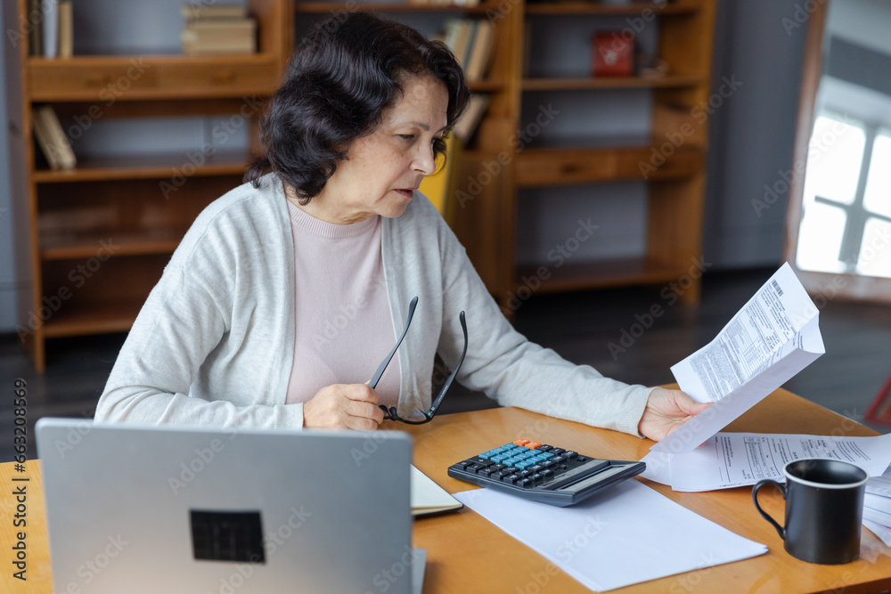 Middle aged senior woman sit with laptop and paper document. Pensive older mature lady reading paper bill pay online at home managing bank finances calculating taxes planning loan debt pension payment