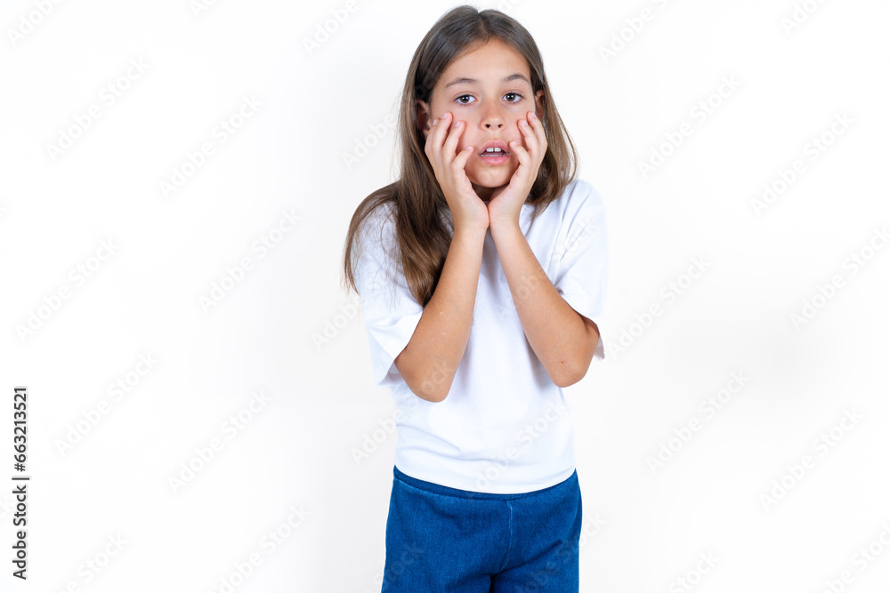 Stupefied Beautiful little kid girl wearing white T-shirt expresses excitement and thrill, keeps jaw dropped, hands on cheeks, has eyes popped out Stupefied Beautiful little kid girl wearing white T-shirt expresses excitement and thrill, keeps jaw dropped, hands on cheeks, has eyes popped out