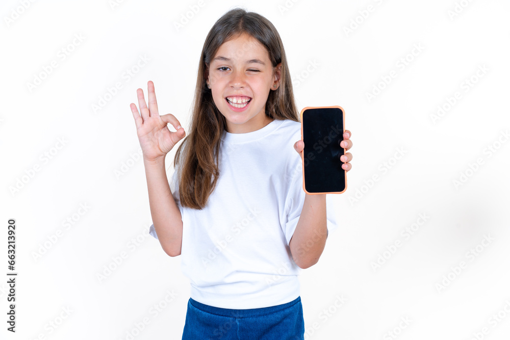 Beautiful kid girl wearing white T-shirt holding in hands cell showing ok-sign Beautiful kid girl wearing white T-shirt holding in hands cell showing ok-sign