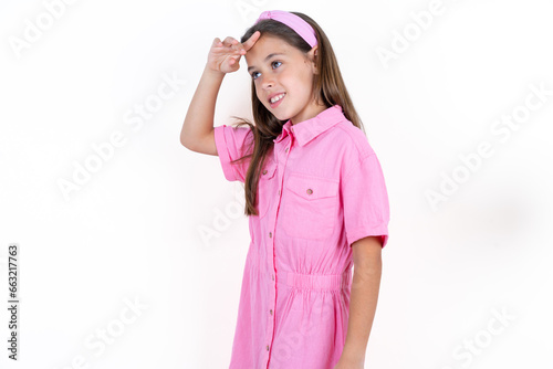 Beautiful little kid girl wearing pink dress very happy and smiling looking far away with hand over head. Searching concept.