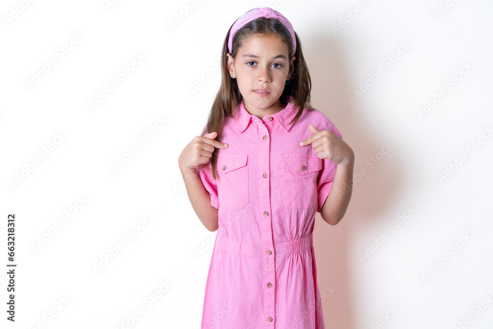 Beautiful kid girl wearing pink dress points at his body, being in good mood after going shopping and making successful purchases Beautiful kid girl wearing pink dress points at his body, being in good mood after going shopping and making successful purchases