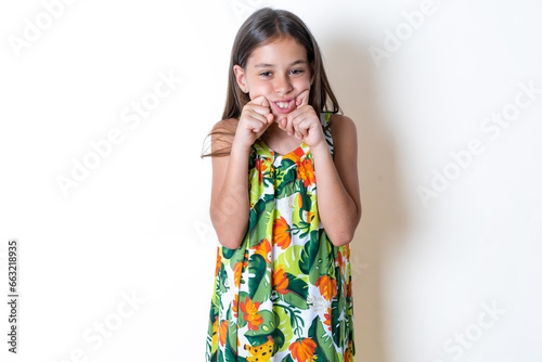 Pleased Beautiful kid girl wearing floral dress with closed eyes keeps hands near cheeks and smiles tenderly imagines something very pleasant