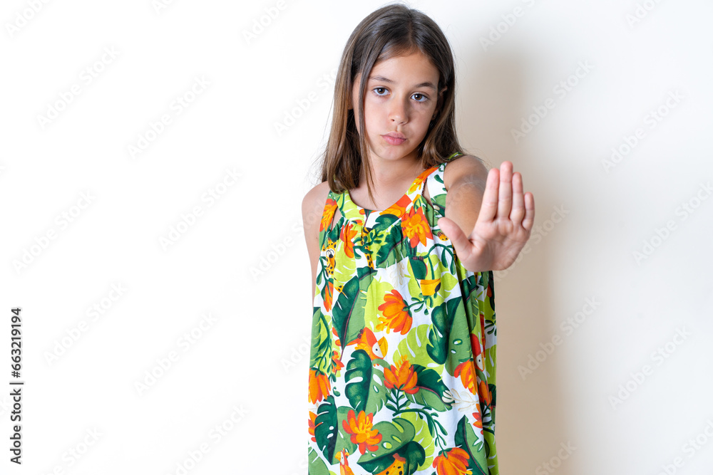 Beautiful kid girl wearing floral dress shows stop sign prohibition symbol keeps palm forward to camera with strict expression Beautiful kid girl wearing floral dress shows stop sign prohibition symbol keeps palm forward to camera with strict expression