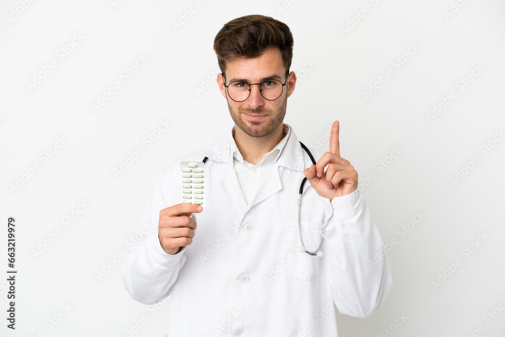 Young doctor caucasian man over isolated on white background wearing a doctor gown and holding pills Young doctor caucasian man over isolated on white background wearing a doctor gown and holding pills