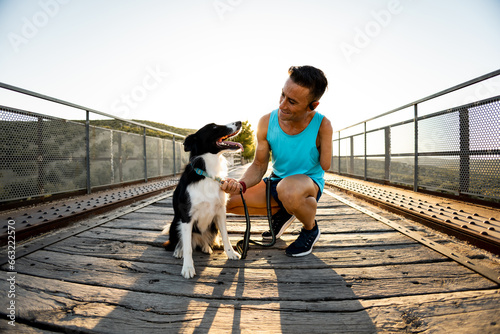An adult man with an amputated arm is crouched down attaching a harness to his dog on an outdoor bridge. Canicross concept, running with a border collie. Sports activities with animals.