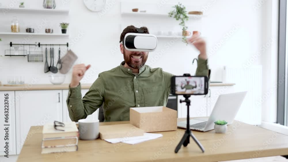 Joyful bearded man wearing VR headset and gesturing from happiness while recording new vlog on modern smartphone. Caucasian technology blogger satisfied with new modern gadget. Joyful bearded man wearing VR headset and gesturing from happiness while recording new vlog on modern smartphone. Caucasian technology blogger satisfied with new modern gadget.