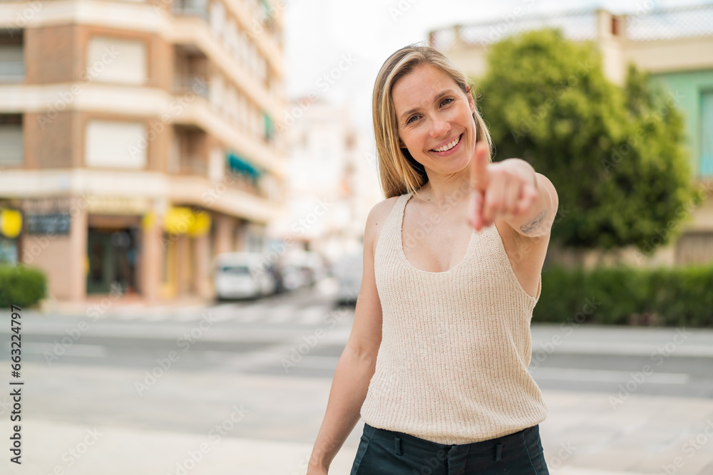 Young blonde woman at outdoors points finger at you with a confident expression