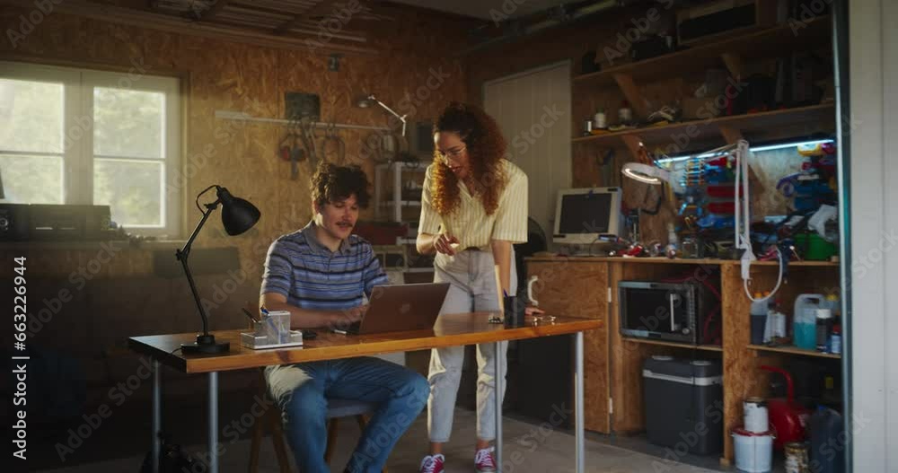 Caucasian Male Software Engineer And Hispanic Female User Interface Designer Talking And Using Laptop Computer in Garage At Home. Diverse Young Startup Founders Working On Innovative Online Service. Caucasian Male Software Engineer And Hispanic Female User Interface Designer Talking And Using Laptop Computer in Garage At Home. Diverse Young Startup Founders Working On Innovative Online Service.
