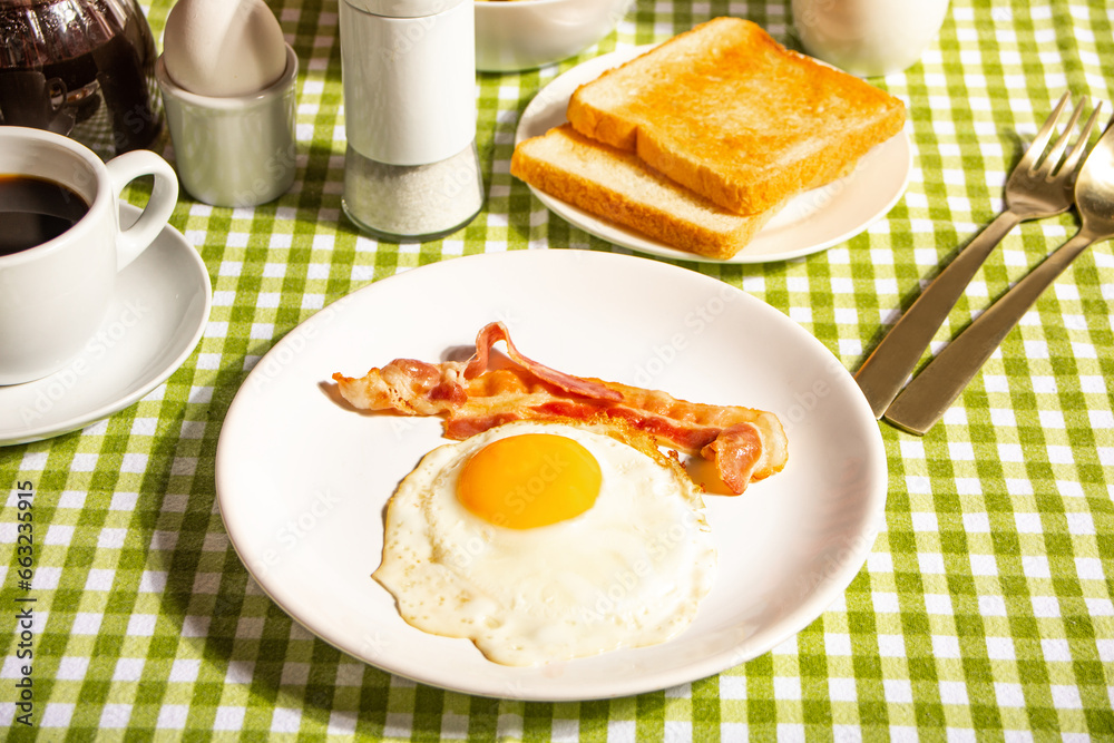 Fried egg with roasted bacon, black coffee, toasts on checkered tablecloth. Retro American breakfast. Hard shadow. Fried egg with roasted bacon, black coffee, toasts on checkered tablecloth. Retro American breakfast. Hard shadow.