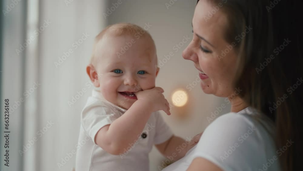 Portrait of happy family affectionate positive mom kissing baby holding in hands looking at window. Mother kisses kid at home. Motherhood, parenting, babycare, childhood, babyhood, love care concept. Portrait of happy family affectionate positive mom kissing baby holding in hands looking at window. Mother kisses kid at home. Motherhood, parenting, babycare, childhood, babyhood, love care concept.