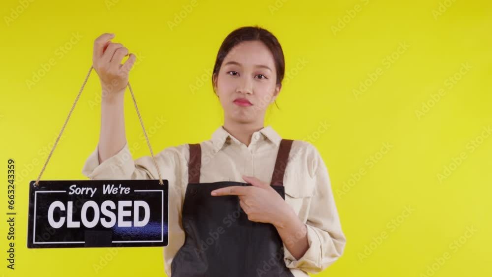 Portrait young asian barista woman wearing apron holding signboard close store with unhappy on yellow background, waitress or entrepreneur showing sign close with stress, small business or startup. Portrait young asian barista woman wearing apron holding signboard close store with unhappy on yellow background, waitress or entrepreneur showing sign close with stress, small business or startup.