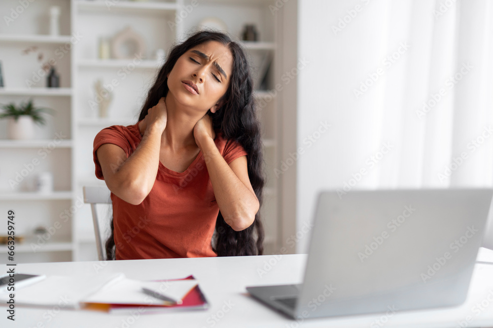 Tired young eastern businesswoman sitting at workplace, rubbing neck Tired young eastern businesswoman sitting at workplace, rubbing neck