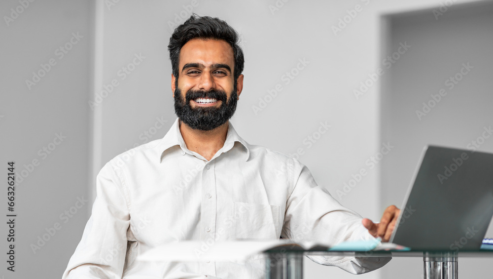 Portrait of happy indian man sitting at desk in office, working on laptop and smiling at camera, panorama, free space Portrait of happy indian man sitting at desk in office, working on laptop and smiling at camera, panorama, free space