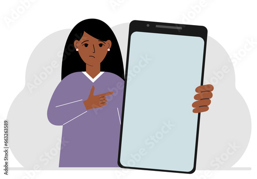 A woman holds a mock-up of a large smartphone with a blank screen and with his second hand points to the screen of the phone.