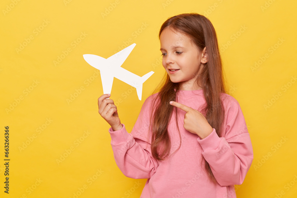 Brown haired little girl wearing pink sweatshirt pointing white paper plane isolated over yellow background advertising airline showing direction copy space for promotions or advertisement. Brown haired little girl wearing pink sweatshirt pointing white paper plane isolated over yellow background advertising airline showing direction copy space for promotions or advertisement.