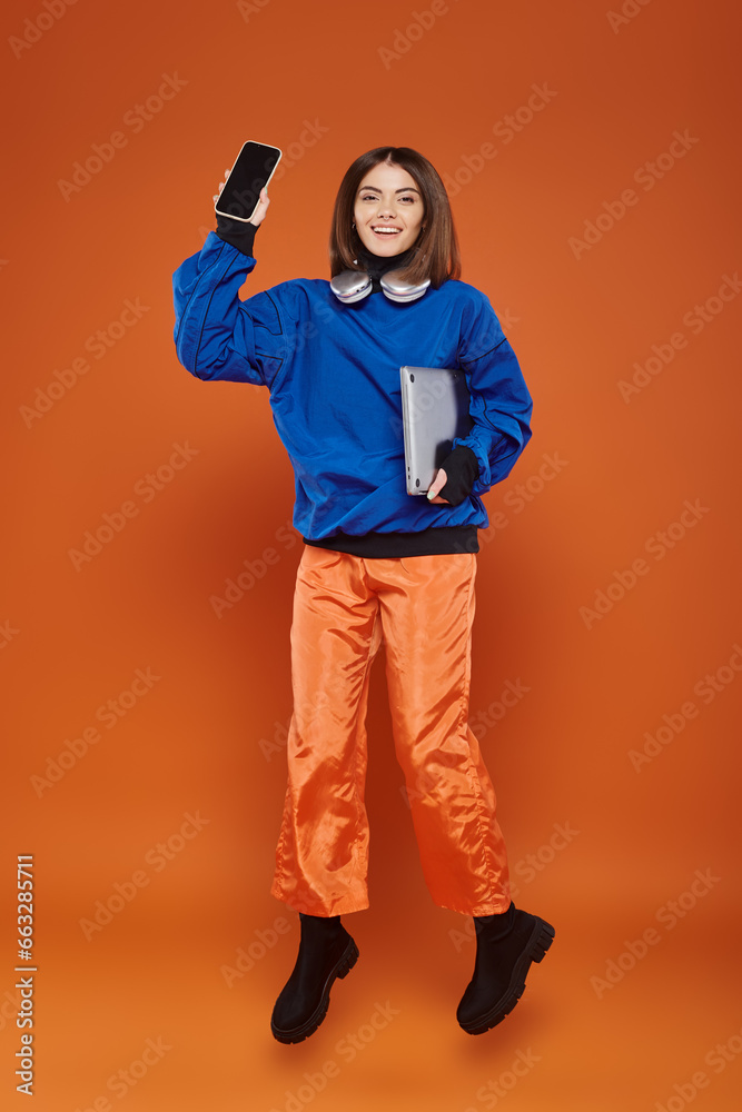 Fototapeta premium cheerful woman in trendy autumnal outfit jumping with gadgets on orange, cyber monday concept