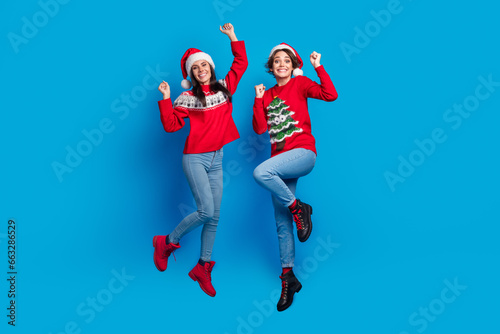Full length photo of cheerful lucky ladies santa helpers dressed print sweaters jumping high celebrating xmas isolated blue color background