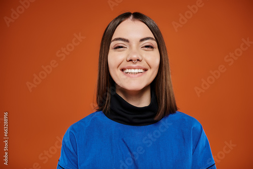 happy young woman with pierced nose looking at camera and smiling on orange backdrop, blue jacket © LIGHTFIELD STUDIOS