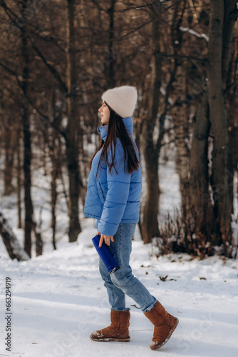 Portrait of young woman in warm clothes in winter forest. Drinking hot tea outdoors from thermos. Hiking.