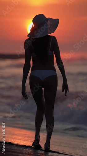 Rear view of silhouette of barefoot slim woman in bikini and straw sun hat walking on sandy beach at sunrise on background of solar disk rising above horizon. Slow motion, handheld shot vertical video