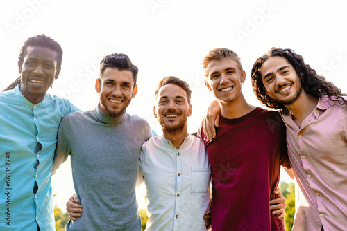A multiracial group of five male friends embraces outdoors, sharing a moment of unity and joy. They all smile warmly, directing their cheerful expressions towards the camera.