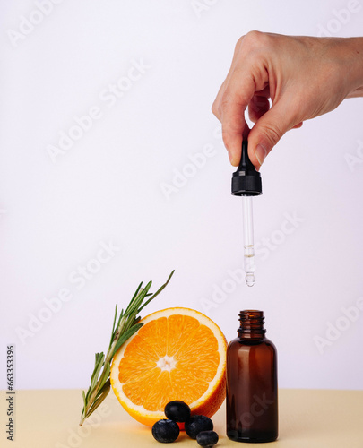 A woman's hand holds a pipette with natural oil cosmetics for face skin and body on the table there is a cosmetic bottle made of brown glass with an orange and a branch of rosemary