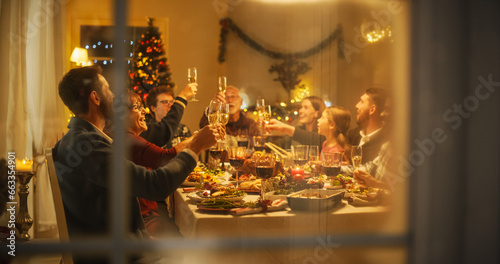 Cinematic Shot Through a Snowy House Window: Christmas Dinner Together with Parents, Children and Friends at Home. Multicultural Family Raising Champagne Glasses and Toasting, Celebrating Holidays photo