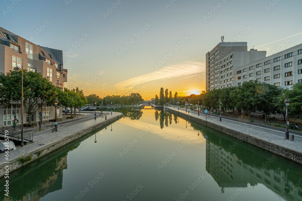 Paris, France - 08 15 2021: Ourcq Canal. Reflections on the Ourcq canal of a bridge, trees, barges and buildings at sunrise.