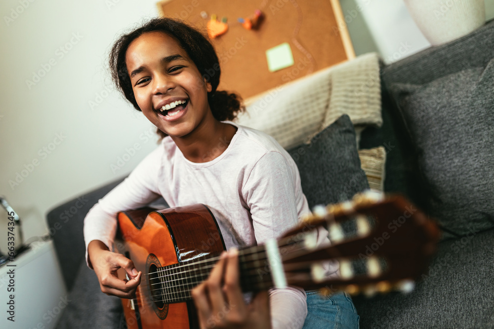 © Mediteraneo - African american teenage girl sitting on couch in her room and learning to play guitar