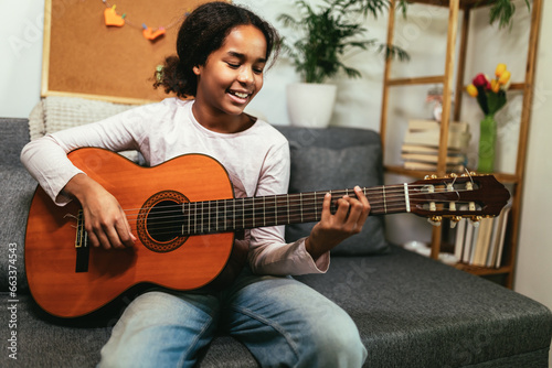 African american teenage girl sitting on couch in her room and learning to play guitar