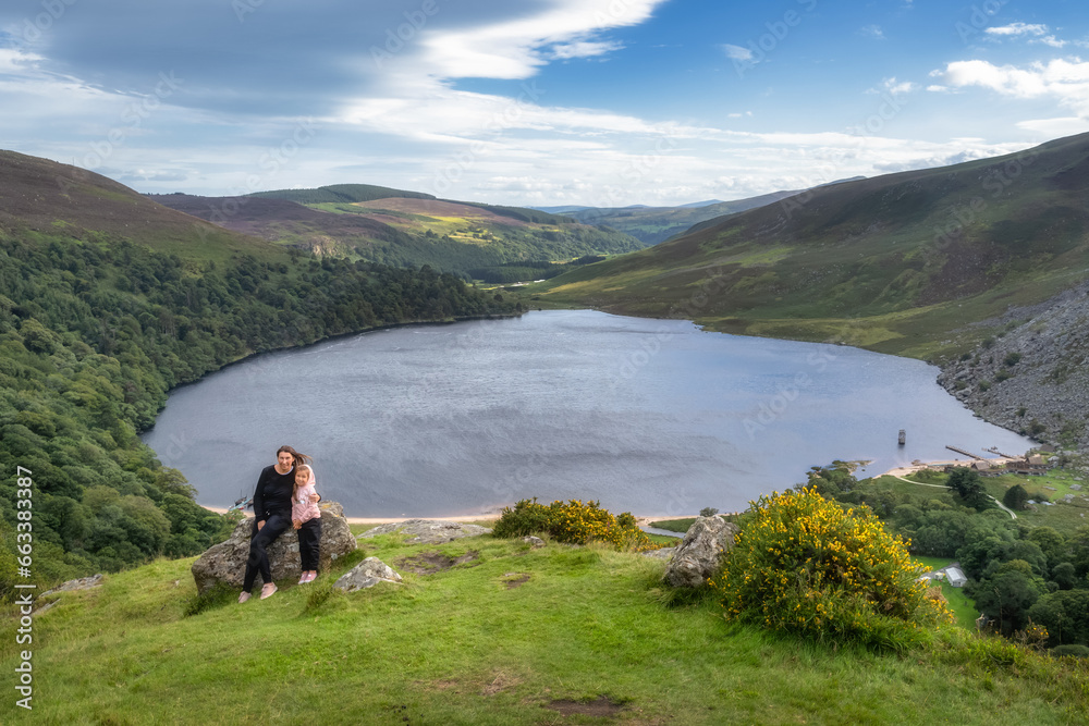 Mother and daughter, family, sitting on a large rock on the edge of cliff. Lough Tay, called Guiness Lake in Wicklow Mountains, Ireland