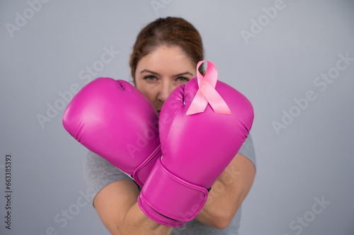 Caucasian woman in pink boxing gloves with a pink ribbon on her chest on a gray background. Fight against breast cancer. 