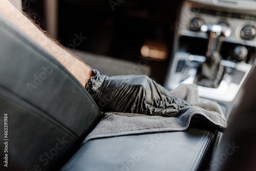 Man's hand in black glove cleaning car interior, dashboard and leather seats with microfiber cloth. Hand wipe down suede leather seat saloon interior. © vladdeep