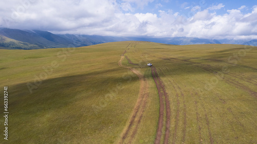 A white SUV is driving in the mountains through the fields. Aerial view from a drone of huge white clouds  snowy mountain peaks and green fields. Dirty off-road with puddles. Splashes from the wheels