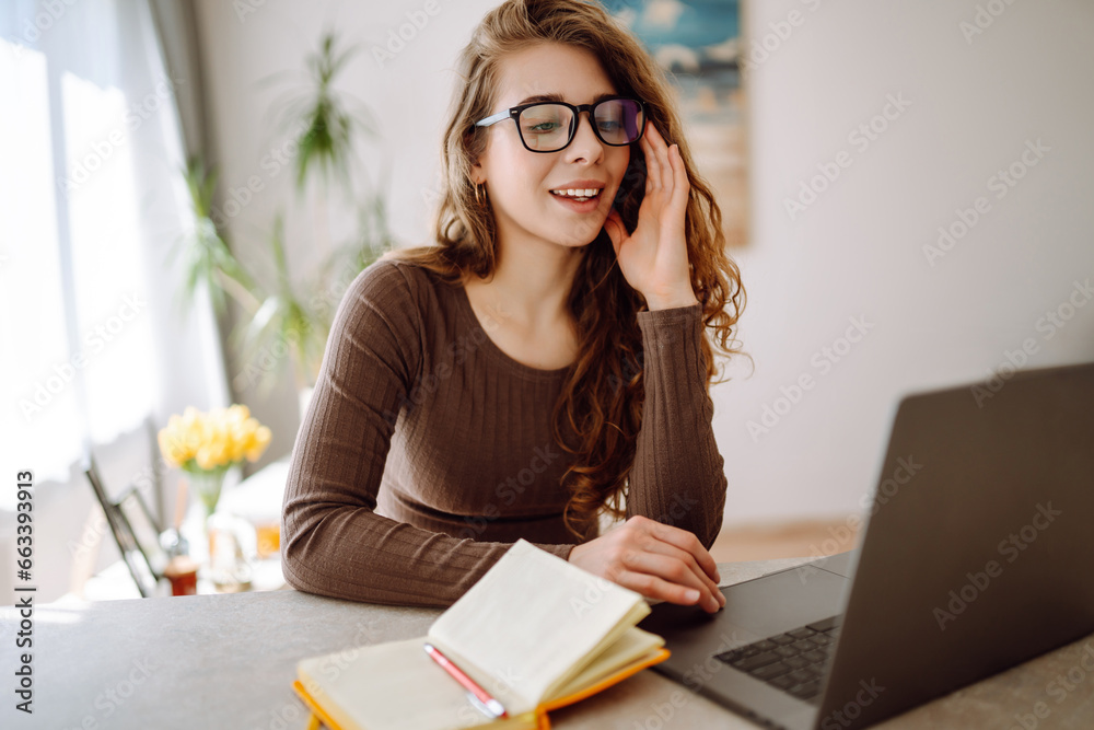 Focused young woman sits at a table with a laptop and a bright notepad, works, looks at the screen, writes at the table. Concept of freelancing, vacation, education. Focused young woman sits at a table with a laptop and a bright notepad, works, looks at the screen, writes at the table. Concept of freelancing, vacation, education.
