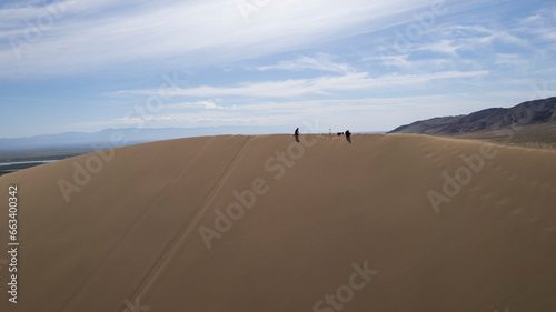 A large sand dune in the middle of the steppe. Drone view of a huge pile of sand. Tourists are walking  enjoying the view. A river runs in the distance and grass grows. The sky with white clouds