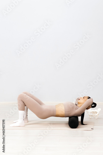 Attractive slim girl using a roller to relax her body muscles after a workout. A young fit woman in light sportswear posing against a light background.