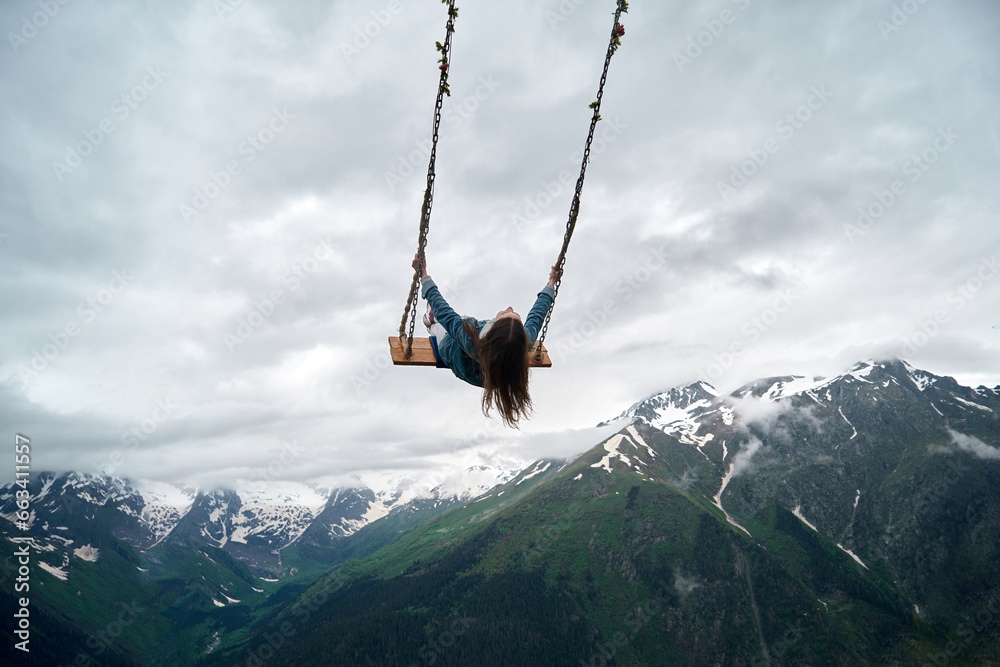 A young woman rides on a swing against the backdrop of snowy rocky mountains. Extreme entertainment in the mountains of the Caucasus