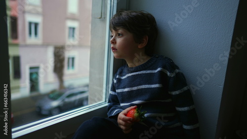 Bored sad emotion of little boy sitting by window staring at view in melancholy, child wanting to go out stuck at home photo