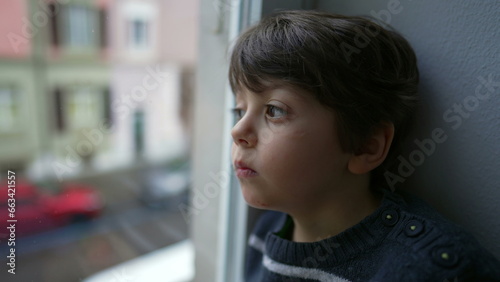 Sad and Bored Little Boy Sitting by Window, Staring in Melancholy, Wanting to Go Out but Stuck at Home © Marco