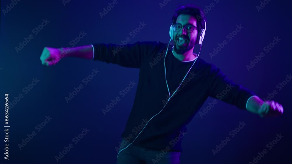 Happy carefree joyful active silly guy having fun moving dancing in club neon ultraviolet background Arabian man dance listening music headphones Indian Muslim male funny move listen audio song party