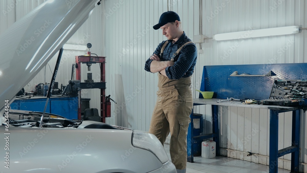 An auto mechanic stands with his arms folded in front of the open hood of a car. The mechanic looks under the hood of the car