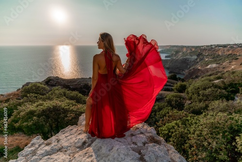 Woman sunset sea red dress, back view a happy beautiful sensual woman in a red long dress posing on a rock high above the sea on sunset.