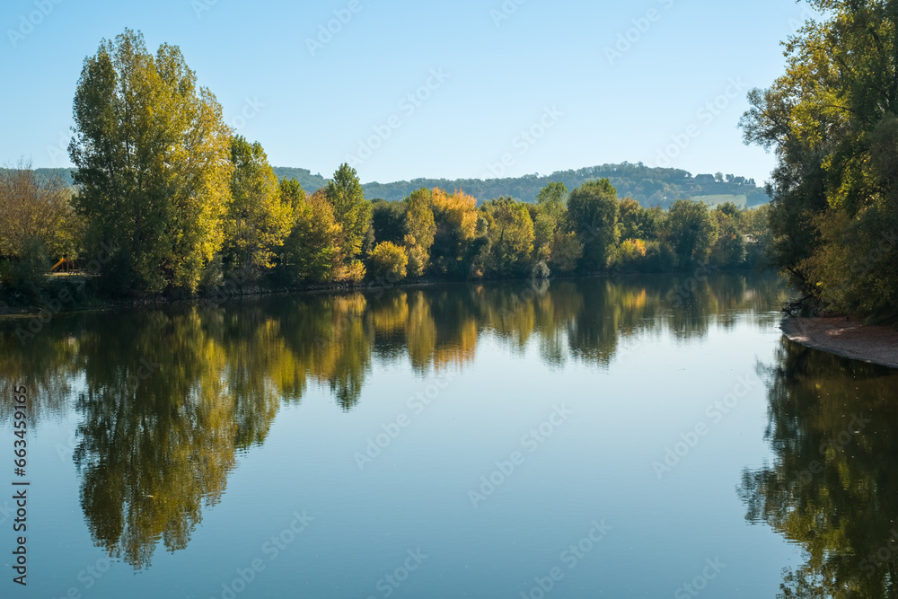 The tree lined banks of the Dordogne river in autumn colours reflected in the mirror calm water at Beynac-et-Cazenac in France