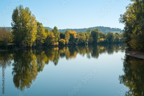 The tree lined banks of the Dordogne river in autumn colours reflected in the mirror calm water at Beynac-et-Cazenac in France