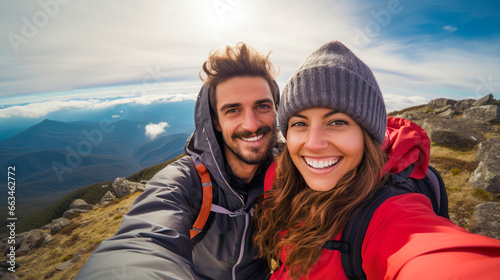 Stylish joyful pair of hikers with backpacks takes selfie at mountain summit, smiling at the camera, on background of large river and peaks
