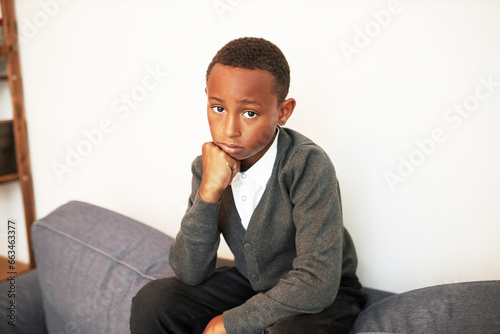 Картината върху платно Upset sad kid boy in uniform sitting on back of sofa with hand under chin lookin